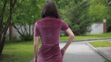 Zarif, açık pembe elbiseli, kendine güveni tam bir kadın. Şehir parkının bulanık ortamında çimlerde tek başına dikilirken kameraya bakıyor ve saçlarına dokunuyor.