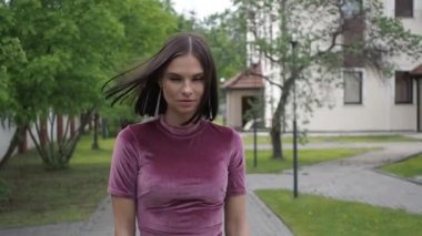 Zarif, açık pembe elbiseli, kendine güveni tam bir kadın. Şehir parkının bulanık ortamında çimlerde tek başına dikilirken kameraya bakıyor ve saçlarına dokunuyor.