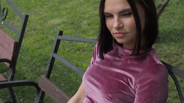 Elegant ung brunett avkopplande på bänk i parken — Stockvideo