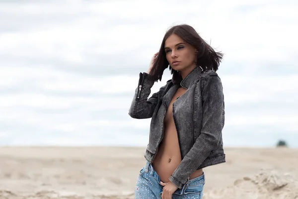 Attractive female model in jeans looking at camera and covering breast with denim jacket on blurred background of countryside