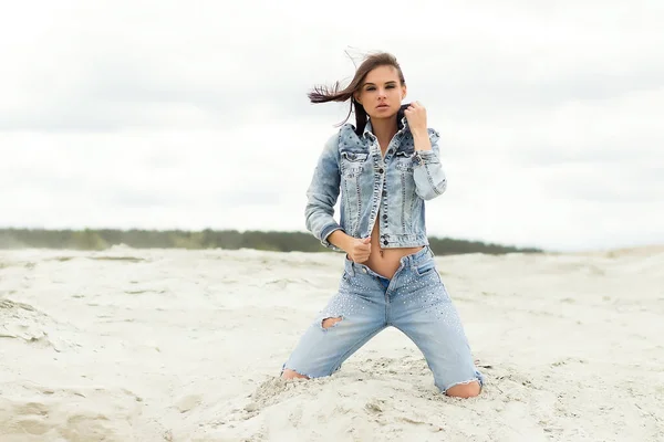 Attractive female model in jeans looking at camera and covering breast with denim jacket on blurred background of countryside