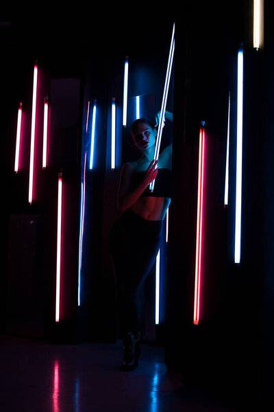 Полное тело чувственная женщина с яркой лампой опираясь на стену в темной комнате с неоновой подсветкой