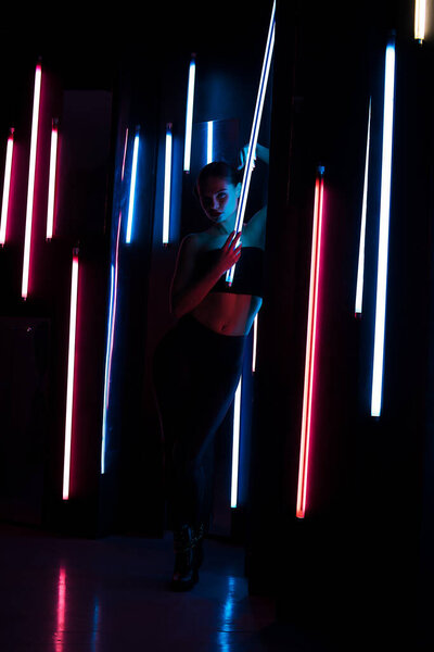 Полное тело чувственная женщина с яркой лампой опираясь на стену в темной комнате с неоновой подсветкой