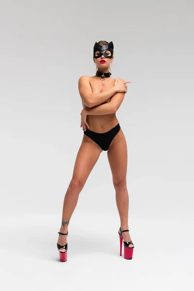 Full body unrecognizable young female with perfect tanned slim body and black mask on face wearing black lingerie and high heeled shoes covering naked breast