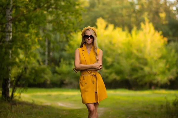 夏に緑の公園のぼやけた背景に立っている間 流行の黄色の衣装を身に着けて腕を横断し カメラを見てスリム女性 — ストック写真