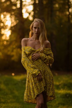 Çiçekli elbiseli sarışın kadın, akşamları ormanda çimlerin üzerinde dikilirken gözlerini kaçırıyor.