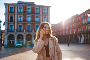 Trençkotlu bir kadın Avrupa şehir meydanında akıllı bir telefonla konuşurken gülümsüyor.