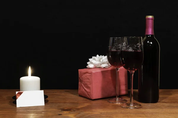 Beautiful etched wine glasses and bottle of red wine, white candle, wrapped present with bow on wooden table with name tag on dark background. Valentines, Mothers Day, Easter, Christmas, Wedding Concepts