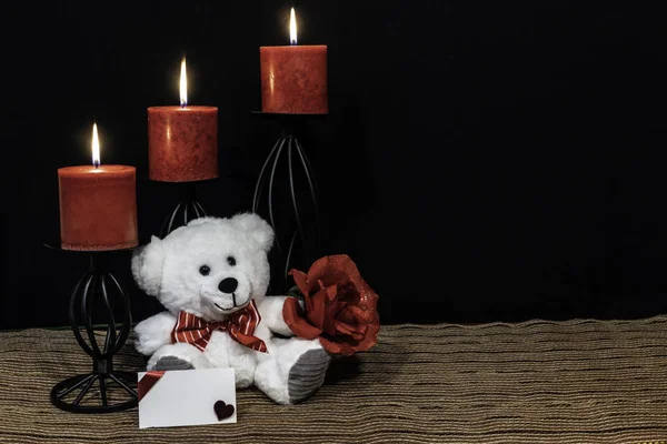 舒适的泰迪熊与红色领结 红玫瑰 红色蜡烛栖息在黑色蜡烛持有人的网状地方垫和木桌与卡和黑暗的背景 情人节 母亲节 复活节 圣诞节 婚礼概念 — 图库照片