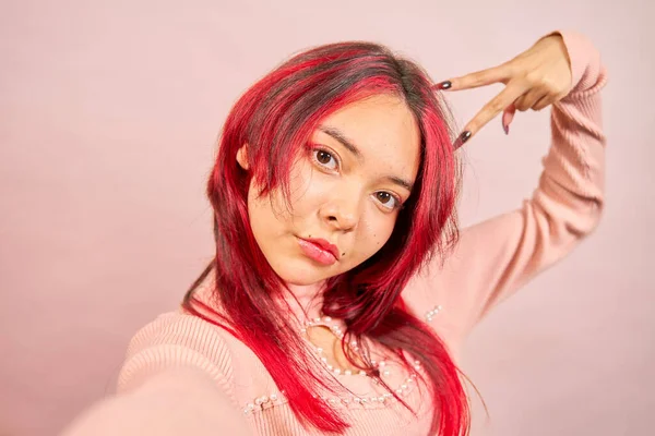 Kızıl saçlı bir genç, pembe arka planda pembe elbiseler giyerek barış işareti yaparken selfie çekiyor.