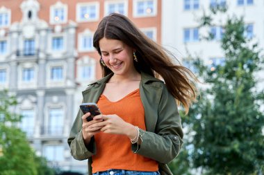 Şehirde yürürken cep telefonu kullanan mutlu genç kadın, internet iletişimi ve kentsel yaşam tarzının keyfini çıkarıyor