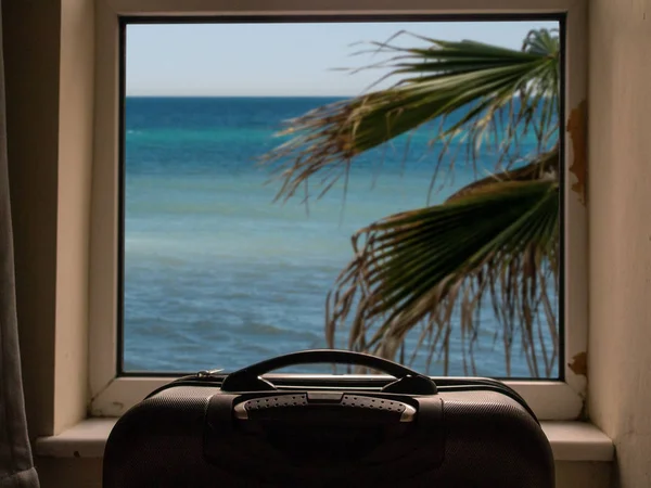 Travel case by a dirty window with view on blue water and palm tree, selective focus, Cheap budget holiday concept,