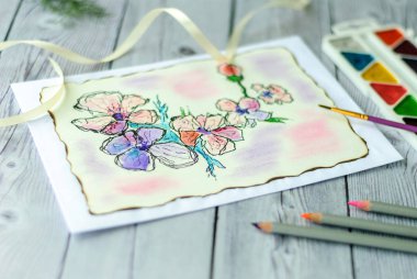 Boş tebrik kartı, el işi zarfı, bebek nefesi çiçekleri, ipek kurdele. Gri ahşap masa arka planı. Düz yatış, üst görünüm