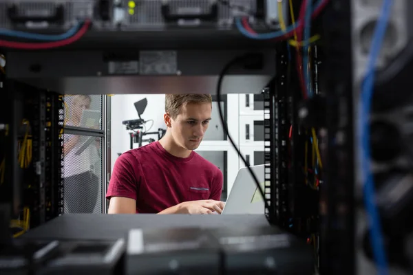 Male Server Engineer Works on a Laptop in Large Data Center.