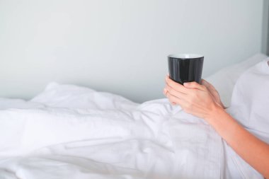 Kadın, kahve bardağını yatağın üstünde tutuyor..