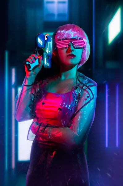 Cyberpunk pink haired woman in neon glasses and transparent raincoat with a handgun on night city background with neon lights.