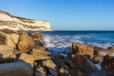 Tranquil view of the Cyprus coastline at evening time with rocks scattered on a sandy beach, calm sea, and colorful sunset sky over the horizon, reflecting the serene beauty of the Mediterranean.