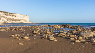 Tranquil view of the Cyprus coastline at evening time with rocks scattered on a sandy beach, calm sea, and colorful sunset sky over the horizon, reflecting the serene beauty of the Mediterranean.