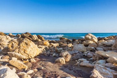 Tranquil view of the Cyprus coastline at evening time with rocks scattered on a sandy beach, calm sea, and colorful sunset sky over the horizon, reflecting the serene beauty of the Mediterranean