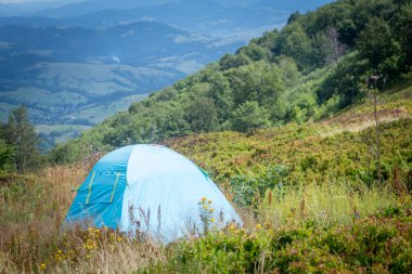  Yaz boyunca Karpatlar 'da yeşil bir çayırda kurulan mavi çadır, dağların manzaralı manzarası, macerayı sembolize ediyor, aktif turizm ve doğada dinlenmeyi.
