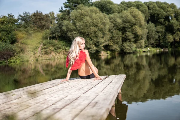 Chica de hadas en Angel Red Wings se sienta en el muelle en la orilla del río. girando suavemente la cabeza — Foto de Stock