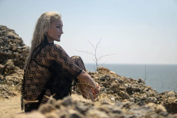 Sommer-Lifestyle-Mode Bild von glücklichen atemberaubenden Frau sitzt auf der Klippe über dem Meer. das Leben genießen und auf das Meer blicken. türkisfarbener Meeresgrund. Stilvolles Kleid, Strohhut, Sonnenbrille — Stockfoto