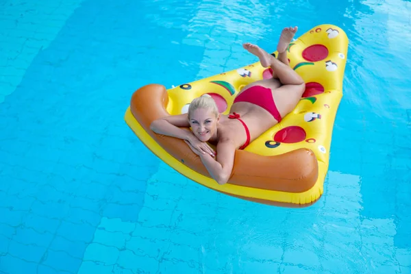 Hermosa chica en un traje de baño rojo flotando en un colchón inflable en forma de pizza en la piscina, hotel turístico —  Fotos de Stock