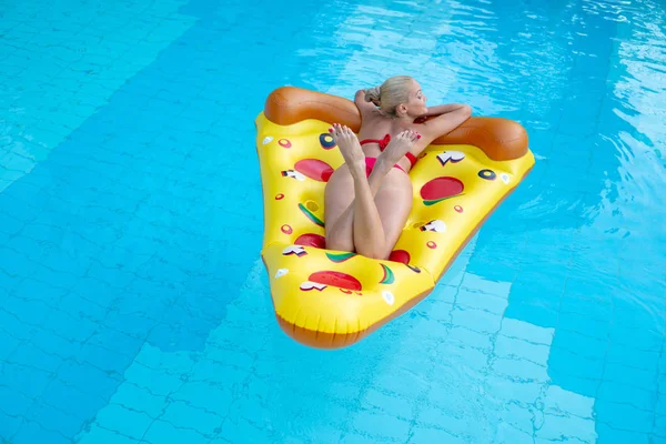 Hermosa chica en traje de baño rojo flotando en un colchón inflable en forma de pizza en la piscina de un hotel turístico —  Fotos de Stock