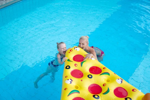 Hermosa familia Mamá y su hija nadan en una piscina de hotel con agua azul y comen un colchón de aire divertido pizza —  Fotos de Stock