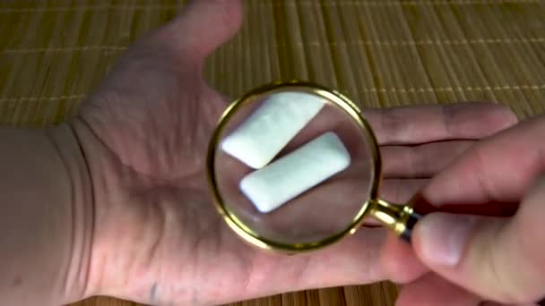 Изучение жвачки под увеличительным стеклом человек с белыми руками две колодки на коричневом фоне, вид сверху — стоковое видео