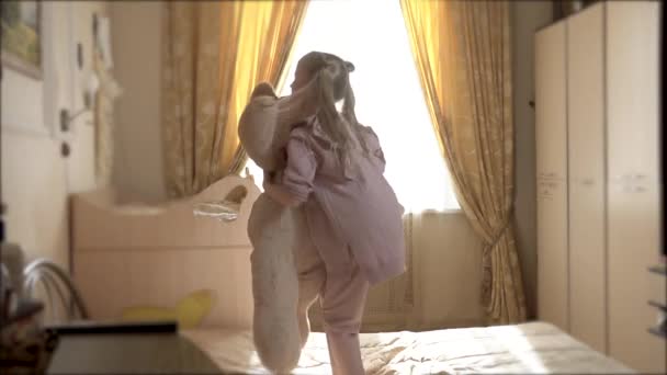 Lustige Teddybär springt auf Bett mit kleinen weißen Mädchen von europäischem Aussehen 8 Jahre alt — Stockvideo