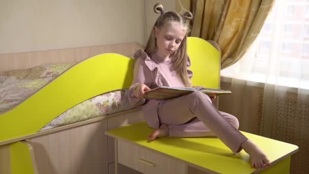 Joyful lit un livre assis sur des meubles amovibles avec un visage joyeux une petite fille dans une chambre d'enfant en vêtements à la mode. Sur le lit et le canapé. — Video