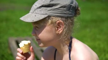 Çömelen kız doğada dondurma yiyor, gri şapkalı yeşil çimlerin üzerinde.