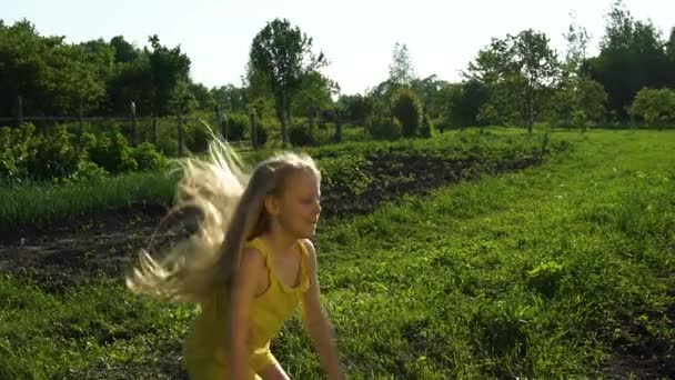 Usmívající se holčička si narovnává vlasy a poskakuje, mladá dívka zírá na kameru a vypadá šťastně, že se jí to líbí na pozadí modré oblohy a zelené trávy na hřišti. Horké léto v přírodě — Stock video