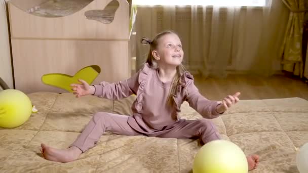 Une petite écolière joyeuse joue avec des boules en gants dans un bel appartement sur un grand lit magnifique avec d'énormes boules et des boules blanches. dans une chambre ensoleillée. — Video