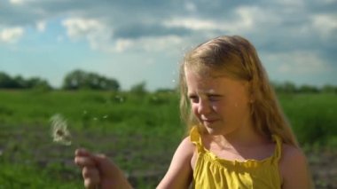 Bir kız yeşil çimen ve köyde uzun sarı saçlı mavi bir gökyüzüne karahindiba üflüyor..