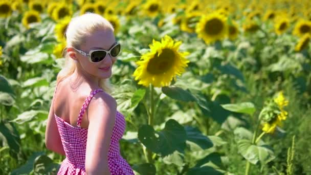 Een meisje in een veld met zonnebloemen in glazen draait zich om en kijkt naar ons, langzaam in een rode jurk vrolijke zomer middag heft glazen — Stockvideo