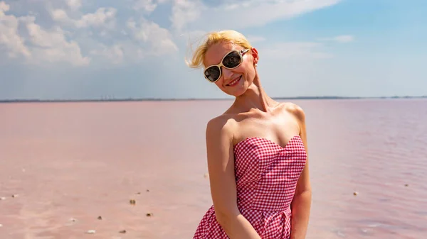 Una ragazza in occhiali da sole si erge contro un mare di acqua salata rosa, in un abito rosso — Foto Stock