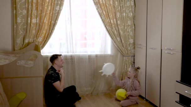 Счастливый молодой отец играет на воздушных шарах в перчатках с красивой маленькой девочкой в детской комнате в домашней одежде. На фоне солнечного окна. — стоковое видео