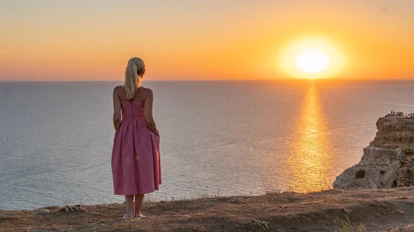 Una alegre chica adulta con una apariencia caucásica mira la puesta de sol con su espalda en el verano por la noche —  Fotos de Stock