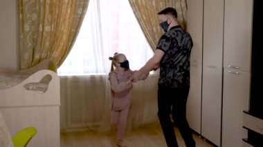 Eğlenen baba, güzel yerli bir bebekle şık kıyafetler içinde çocuk odasında dans ediyor. Güneşli bir pencereye karşı. Maskeliler.