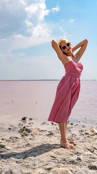 Hermosa chica en un vestido rojo en un lago de sal roja con nubes azules en gafas de verano —  Fotos de Stock