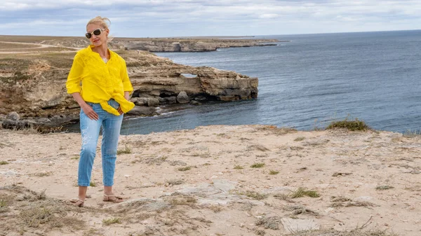 Una ragazza sul bordo dell'abisso sta in jeans e camicia gialla, intorno al mare e la roccia assomiglia a un coccodrillo. — Foto Stock