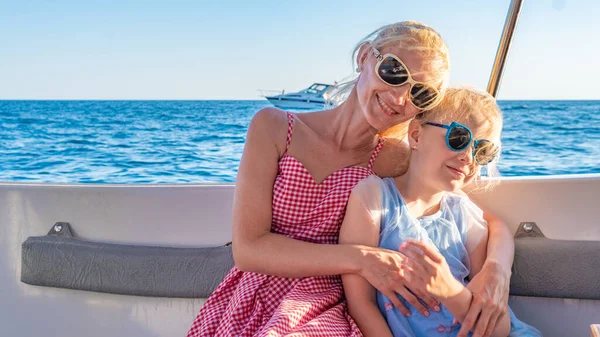 Ragazze gioiose sedersi su uno yacht, giovane con labbra delicate, adulto abbraccia la figlia. Intorno al mare, godere in occhiali da sole in estate — Foto Stock