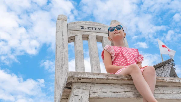 Una bambina si siede con gli occhiali da sole su una sedia di foto qui sotto, estate con nuvole blu — Foto Stock