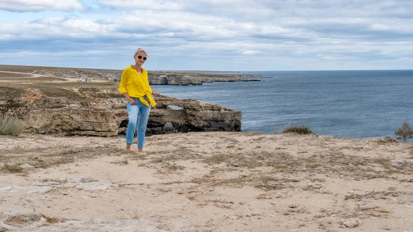 Felice signora in camicia gialla giovane con bellissimi occhi sullo sfondo del mare durante il giorno e in estate su una scogliera — Foto Stock