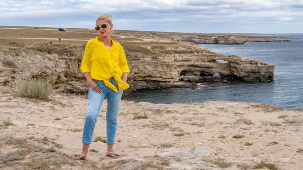 Allegro signora in camicia gialla con uno sguardo gentile sullo sfondo delle montagne e dell'oceano con nuvole blu nel caldo sole su una spiaggia di sabbia — Foto Stock