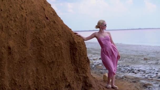 Mujer alegre en un vestido rojo con una apariencia carismática en el fondo de un lago rosa con un cielo azul durante el día y el verano — Vídeos de Stock