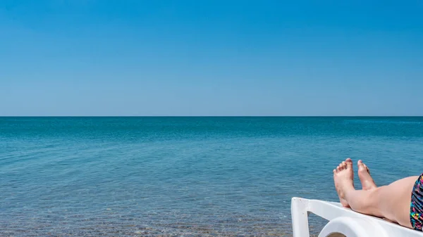 Sdraiato sul mare, acqua blu, piedi su una panchina bianca — Foto Stock