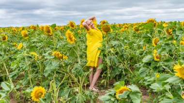 Sarı elbiseli neşeli bir kız, tarlanın arka planında sevimli bir gülümseme, ayçiçekli mavi bulutlar, gün boyunca ve yaz boyunca.